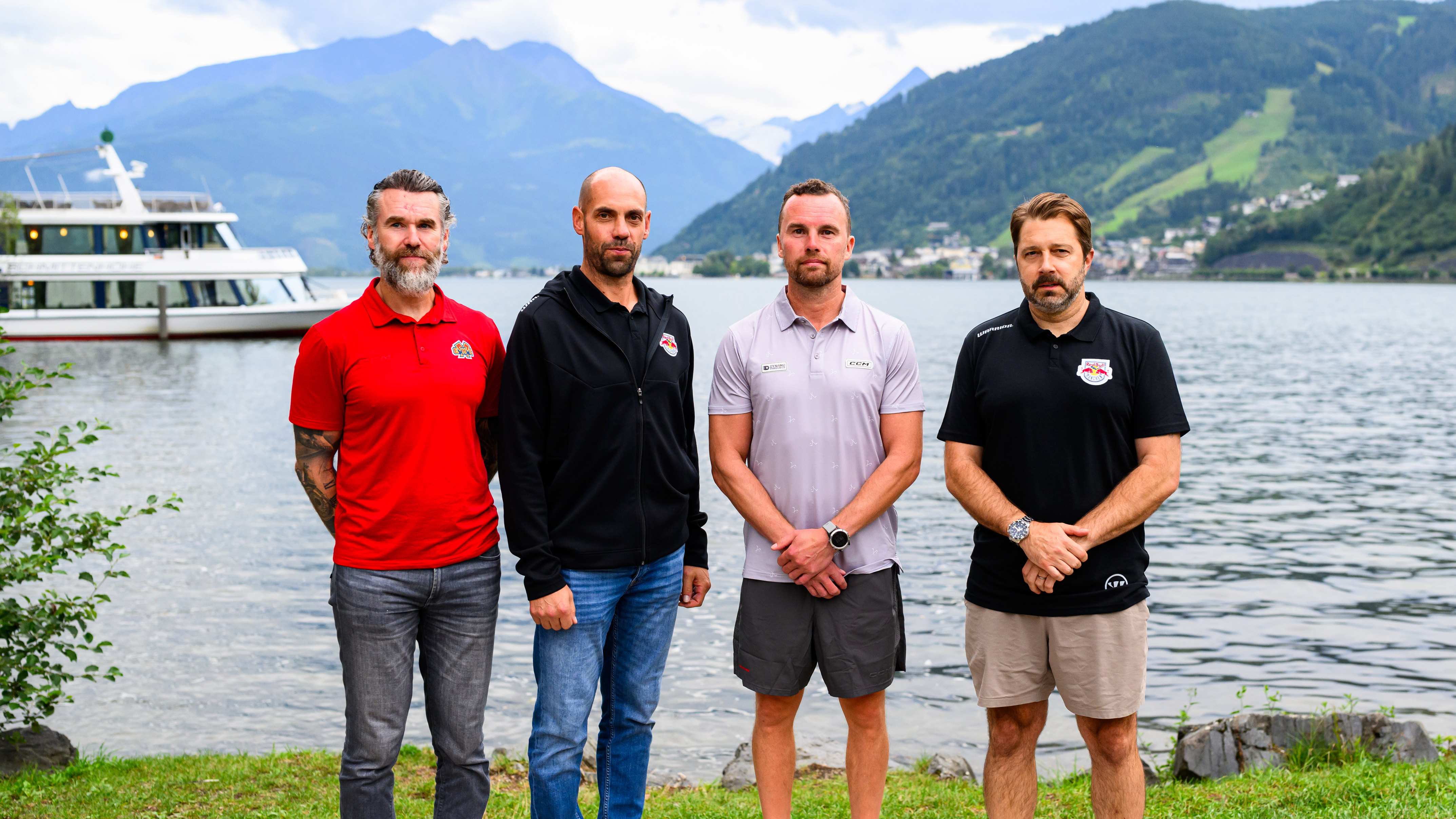 Vier Männer stehen am Ufer eines Sees mit Bergen im Hintergrund.