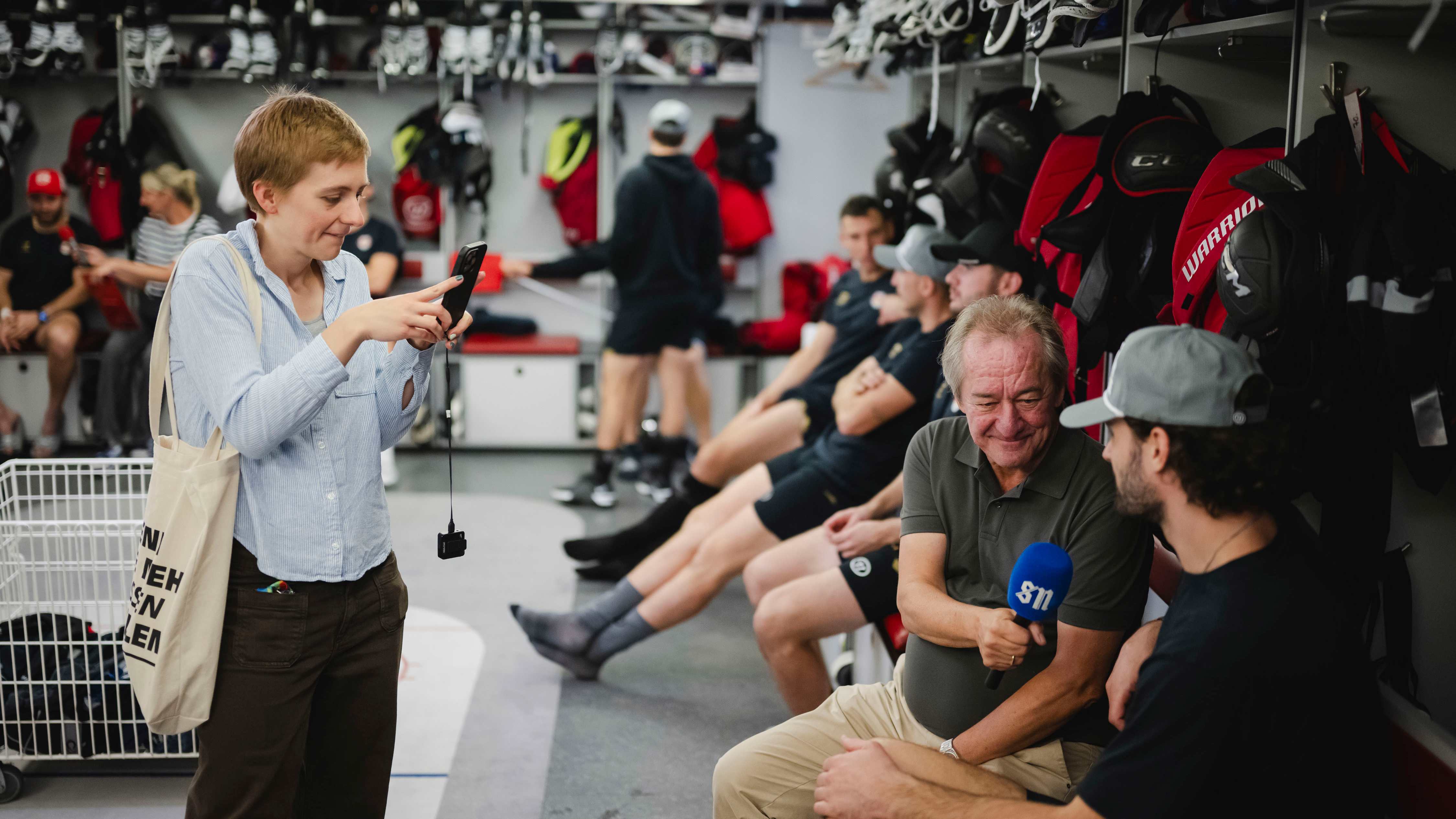 Eishockeykabine: Reporterin filmt mit Handy, Journalist interviewt Spieler
